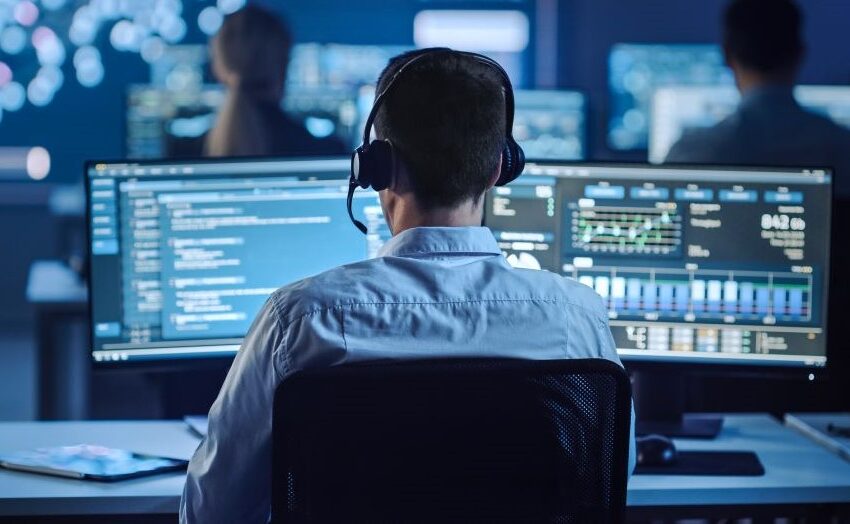A man sat at a computer, wearing a headset, in what looks like a security operations centre